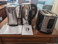 Photo of three stainless steel kitchen appliances including Hamilton Beach electric can opener with manual on the left, Lagostina electric kettle with manual in the center, and Hamilton Beach toaster with buttons and dial on the right. Items placed on a wooden table in a living room setting.