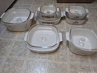 Six vintage CorningWare ceramic dishes with original glass lids viewed from above on tiled surface. They display the Spice of Life floral and spice motifs, all in excellent condition.