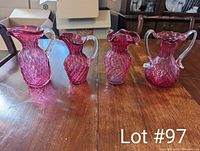 Four vintage cranberry glass pieces including three pitchers and one double-handled vase. All have diamond patterned texture, clear glass handles, and deep red color.