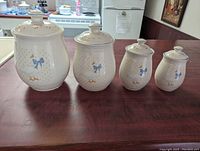 Photo showing four vintage ceramic canisters in graduated sizes with lids, decorated with country geese and ducks with blue bows on dotted background glaze, arranged on wooden table.