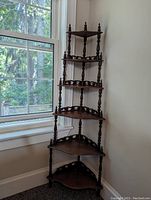 Full view of vintage walnut corner whatnot shelf placed in a corner next to a window with six triangular shelves and turned spindle supports