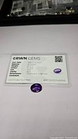 Oval cut deep purple amethyst gemstone placed on a white surface accompanied by a GRWN GEMS certification card detailing weight, color, clarity and dimensions.