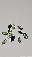 12 marquis cut tourmaline gemstones showing mixed tones of green, blue, and dark green under bright direct light on a white background.