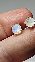 Close-up front view of one rainbow moonstone earring held between fingers showing blue flash and 4-prong 925 sterling silver setting.