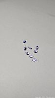 Eight oval cut tanzanite gemstones displayed on a gray surface, showing their oval shapes and varying purple-blue colors.