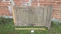 Two antique wooden cupboard doors with raised panels leaning against a brick wall, showing original metal hinges, weathered wood surfaces, and aged patina. A measuring stick is placed in front for scale.