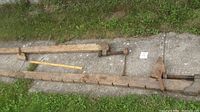 Two large antique wooden clamps laid out on concrete and grass, showing length comparison with measuring stick and notches along wooden body.