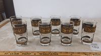 Eight clear glasses arranged in two rows on a cream patterned tablecloth. Glasses feature black and gold bird design band near rim. All glasses shown upright, no chips or cracks visible.