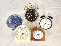 Front view of the lot showing five vintage alarm clocks with various shapes, colors and materials, arranged on white cloth background.
