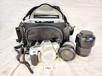 Minolta film camera body with attached lens, additional zoom lens with lens cap, and gray padded camera carrying bag. Lot includes camera equipment and accessories.