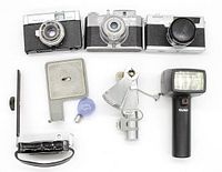 Top view of three vintage cameras with lenses, photographer's flash, wired shutter release and bulb flash arranged on white surface.