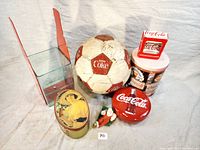 Overview of vintage Coca-Cola items including a soccer ball, tins, coin bank, cigarette case, and figurines.