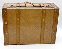Side view of suitcase showing brown patterned faux-leather surface with fleur-de-lis accents, rope handle, leather straps, and metal hardware.