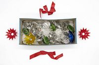 Box containing 11 vintage glass flower-shaped Christmas tree bulb covers in various colors including clear, green, yellow, and blue. Two red metal candle holders with star-shaped candle bases are placed outside the box. Two pieces of red ribbon are also visible.