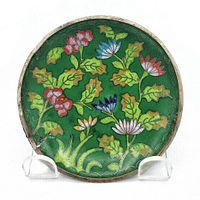 Full front view of the round cloisonné plate showing colorful flower and leaf pattern on green enamel background with metal rim.