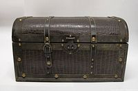 Front view showing faux crocodile leather textured trunk with curved lid, leather straps and metal rivets, central lock and buckle detail.