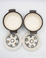 Two open ceramic bowls with black/brown handles showing cream interiors and decorative lids turned upside down in front.