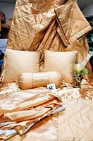 Gold satin quilted comforter folded with two square pillows, one cylindrical pillow, and a clear glass vase with hand-painted floral decoration in front.