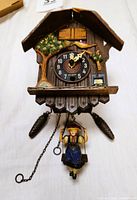 Full front view of the chalet-style wooden cuckoo clock showing the clock face, girl on the swing pendulum, pine cone weights, tree and flower decorations, and visible roof damage.