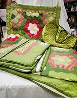 Set includes folded patchwork quilt with hexagon flower patterns in green, red, white, and cream colors, two matching folded shams, and a fabric tote bag with green Christmas tree print and black edges and handles.