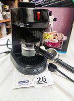 Front-angle view of black DeLonghi Caffe Pronto espresso machine with portafilter attached, black measuring scoop, original packaging and instruction manual in background.