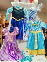 Photo showing four Disney princess dresses hanging: Elsa, Anna, Rapunzel, and Merida, plus turquoise gloves at front.