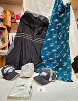 Black zippered jacket, blue-green Eagles sleep pants, three Eagles baseball hats, Eagles notebook in packaging, and ceramic Super Bowl XVIII mug displayed on table