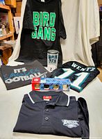 Full lot view showing four shirts with Philadelphia Eagles designs, a set of 4 NFL collector shot glasses in box, and a large green Eagles plastic mug with lid.