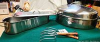 Photo showing the stainless steel high dome roasting pan with handles and lid, Norpro fish poacher, and two vintage meat carving forks with wooden handles on a green surface.