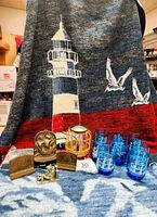 Full view of the nautical themed lot with the Biederlack reversible acrylic throw showing lighthouse and seagulls design, blue rock glasses with sailing ship prints, bronze colored metal bookends featuring nautical ships, pewter captain figurine, and rattan wax melt light.