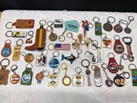 Top-down view of approximately 38 vintage key chains laid out on a white surface, showing the various designs and materials including metal, plastic and leather.