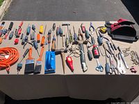 Tools laid out on a table under sunlight showing full view of the lot including screwdrivers, pliers, hammer, paint brushes, tape measure, tool bag, extension cord, and various hand tools.