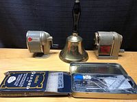 Overview of lot showing two manual pencil sharpeners, a vintage brass bell with handle and Helix Oxford mathematical instrument set with box and tin case.