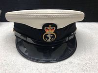 Front view of the navy officer peaked cap showing the gold embroidered crown and anchor insignia on a black patch, white textured top, black band and shiny black visor.
