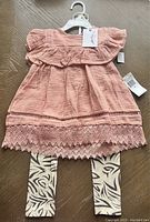 Coral toddler dress with ruffles and lace hem over animal print leggings, on a wooden floor, shown on a hanger with tags.
