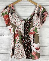 Front view of blouse hanging on a wooden hanger against white wooden background showing floral patterns and cold shoulder sleeves.