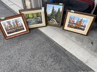 Four framed oil paintings displayed outdoors, showing forest and river landscapes.