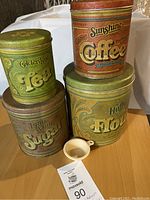 Set of four vintage metal kitchen cannisters labeled Coffee, Tea, Sugar, and Flour with a small white scoop placed on a tabletop.