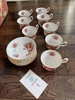 Eight Royal Albert tea cups and eight matching saucers arranged on wooden floor, showing rose floral pattern and gold trim. Cups have some wear and minor chips on rims.