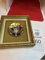 Photo of the framed painted porcelain plate depicting a woman in a blue hat, placed on a white surface with a red background.