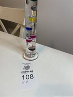 Full view of the tall cylindrical glass Galileo thermometer with colorful floating bulbs on a white table.