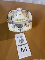 Square ceramic sugar bowl with lid, floral and gold scroll painted decoration, blue trim, photographed from top angle on wooden surface with auction tag