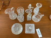 Top-down photo of nine clear vintage crystal pieces including pitchers, candle holders, small bowl, covered trinket box, glasses, and a small bottle on a wooden surface.