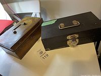 Two vintage lockboxes displayed side by side: a wooden box with an attached metal key and a personalized decoration of newspaper clippings and a metal letter 'K', and a black metal lockbox with latch and handle.