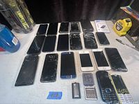 Lot of 19 cell phones with scratches and cracked screens spread on table plus two batteries and a tape measure.