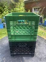 Stacked green and black plastic milk crates shown outdoors with grass and house in background.