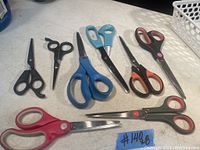 Photo showing 8 pairs of scissors in different sizes and handle colors with metal blades on a light surface.