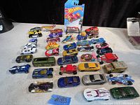 Wide view of 27 Hot Wheels cars on table, mostly loose with one still in sealed showroom packaging. Various models and colors shown with some age wear.
