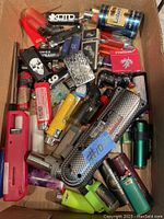 The photo shows a box containing about thirty assorted lighters of various styles, colors, and brands. Included are disposable plastic lighters, metal torch lighters, and some with unique designs and textures, mostly vintage used with some new ones.