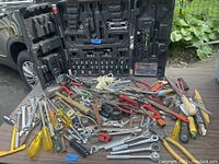 Overview of large assortment of hand tools including those inside and outside of black carrying case, displayed on ground showing normal use and wear
